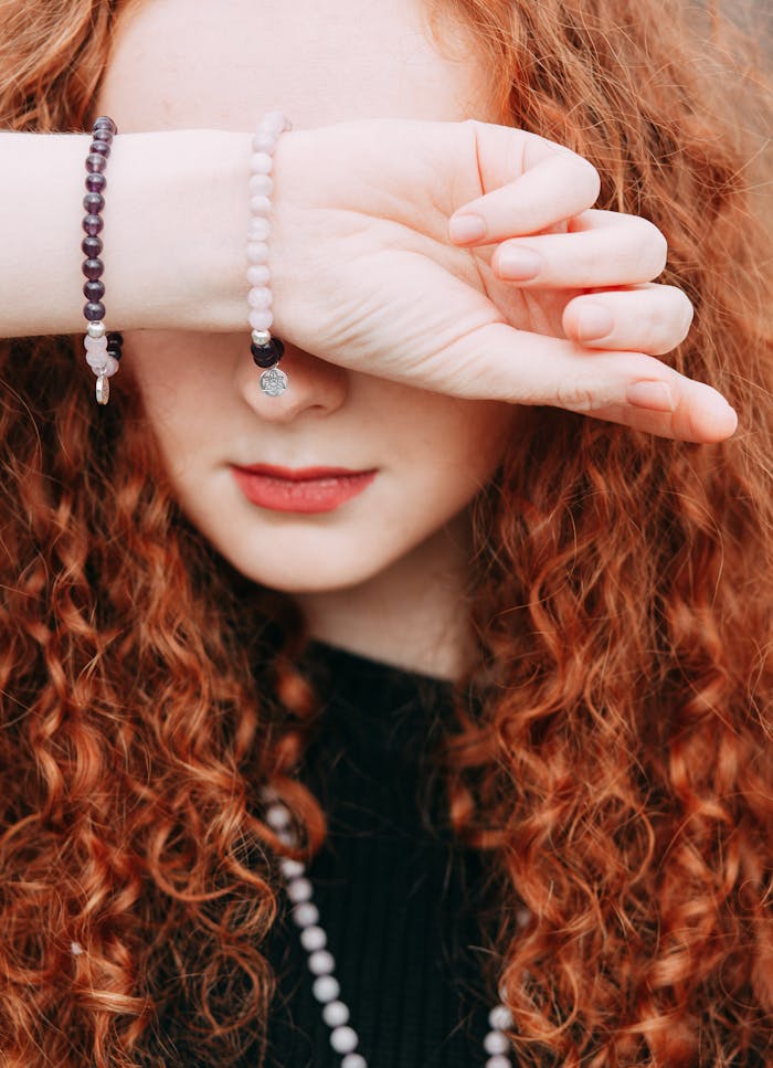 Close-up of a redheaded woman with curly hair covering her eyes, showcasing bracelets.
